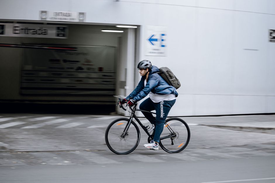 A man in a blue jacket cycling on an urban sidewalk with a backpack.