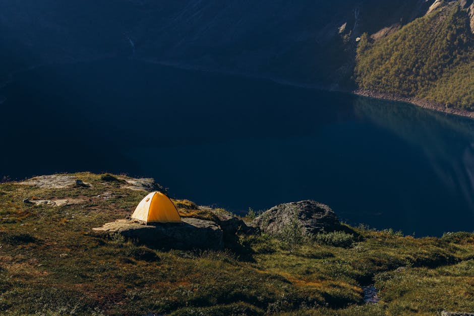 A solitary yellow tent on a grassy hill overlooking a tranquil mountain lake at dawn.