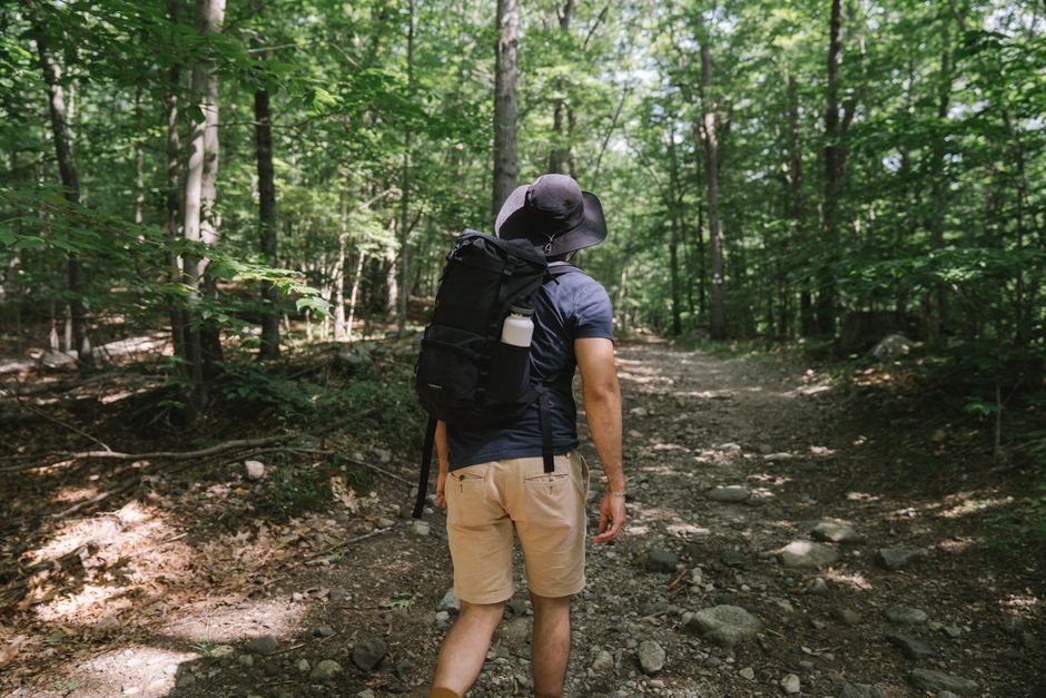 A man walks on a forest trail, wearing a backpack and hat, surrounded by trees.