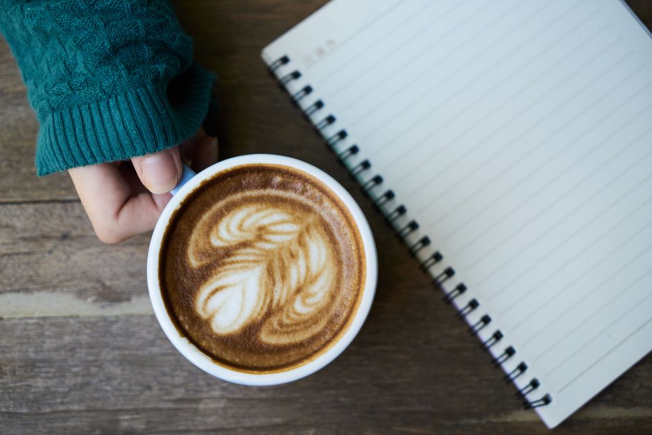 Hand holding a cappuccino with intricate latte art beside a blank notebook, perfect for creative reflection.