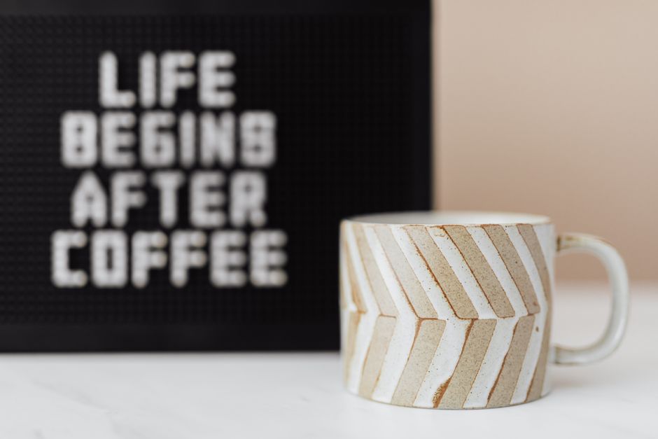 Ceramic coffee cup with creative design placed near blurred peg message board with Life Begins After Coffee message on table near pastel pink wall