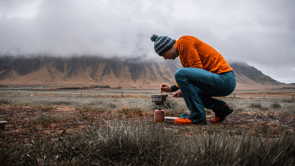 Man cooks with portable stove in scenic Icelandic landscape surrounded by foggy mountains.