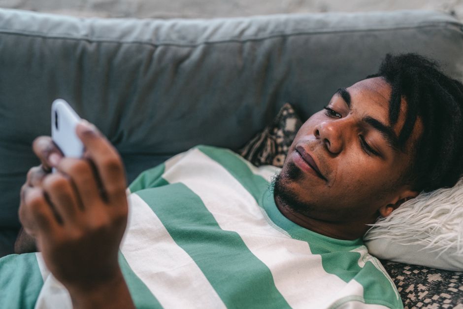 Man with dreadlocks lying on sofa, using smartphone indoors.