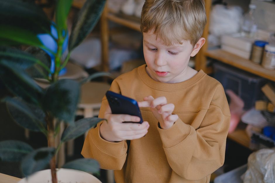 A young boy in a brown sweater uses a smartphone near a potted plant indoors.