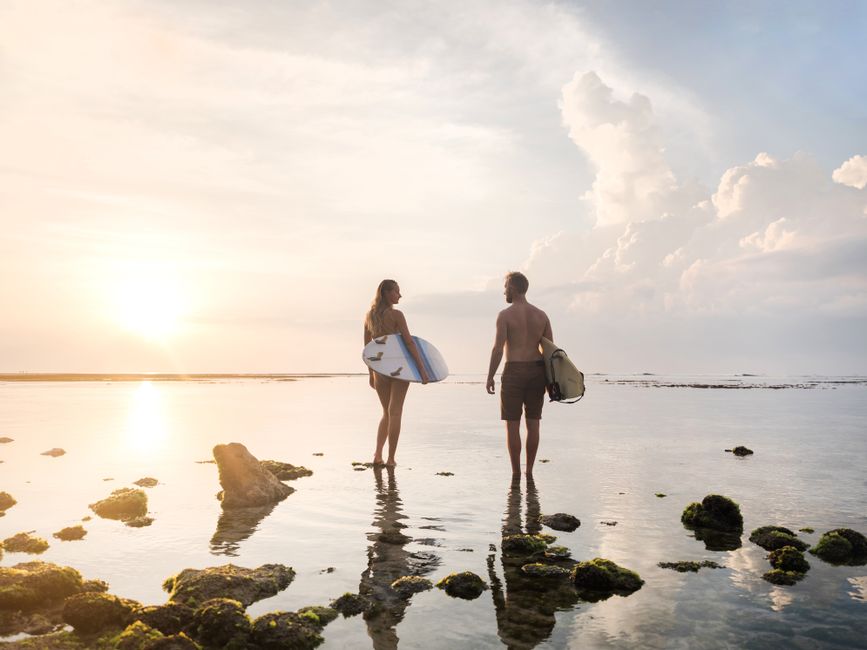 A couple with surfboards standing in shallow ocean water during sunset, enjoying a peaceful beach moment.