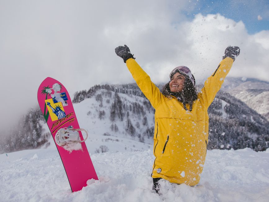 Smiling woman in yellow jacket celebrating snowboarding in winter mountains.
