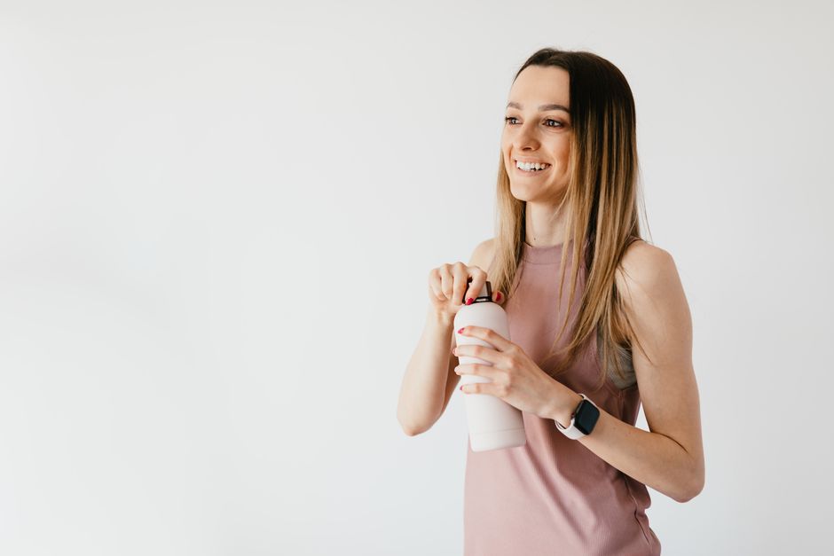 Cheerful smiling young female in casual wear with smart watch looking away while opening cosmetic bottle and standing against white background