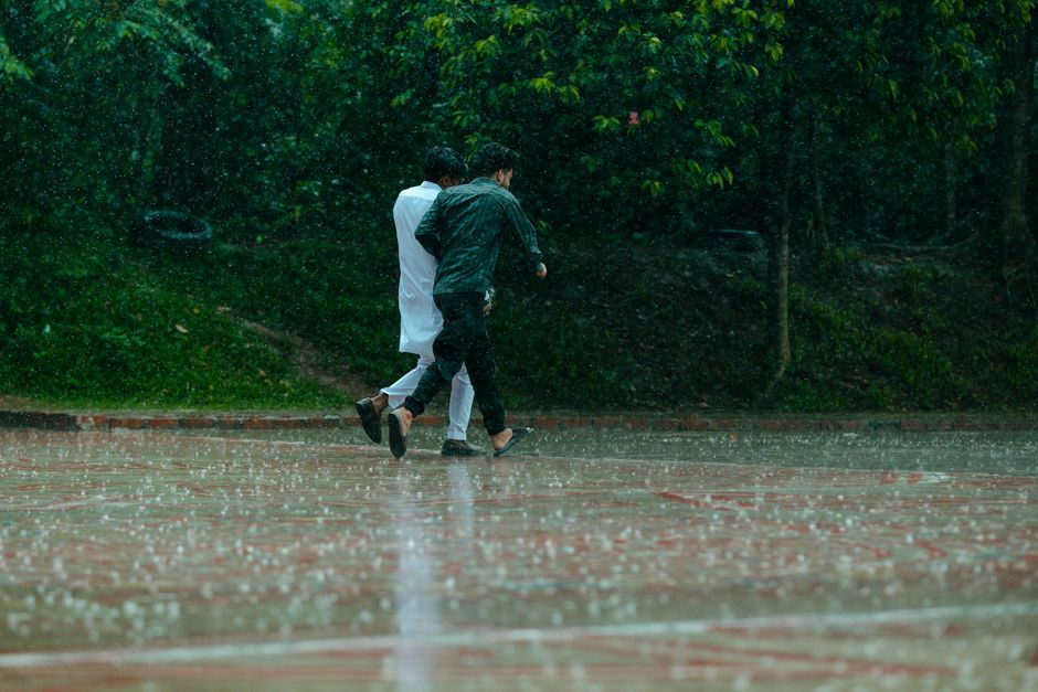 Two people walk together in heavy rain on a paved outdoor path surrounded by greenery.
