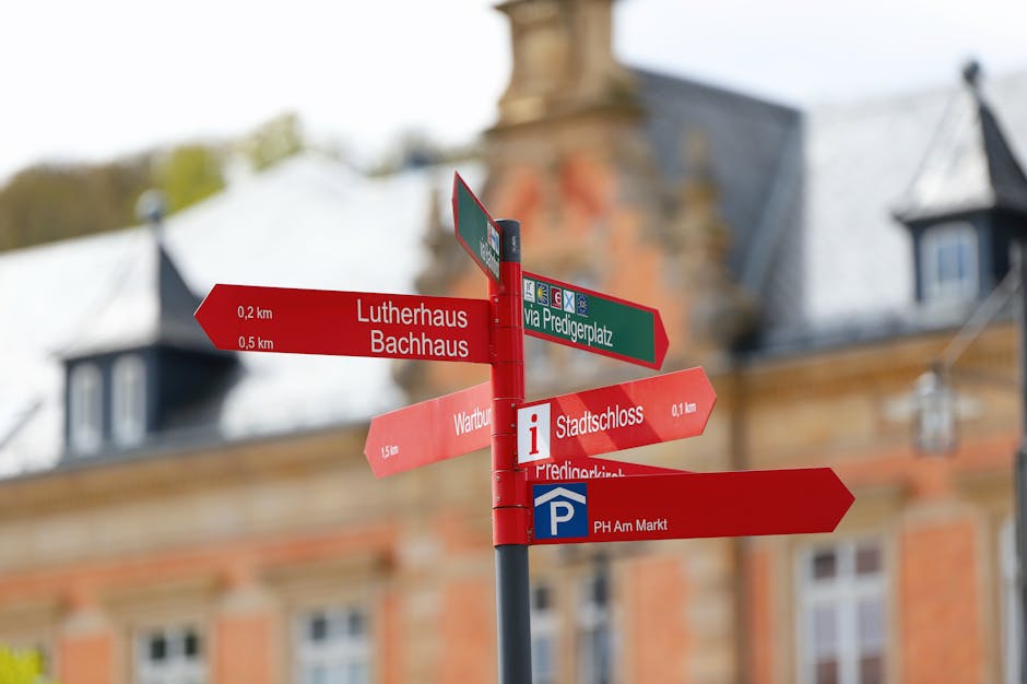 Bright red directional signpost with landmarks against historic European architecture.