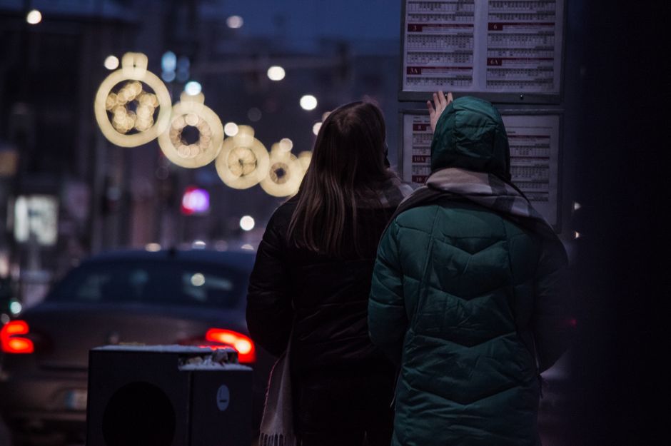 Two people check a bus schedule on a winter night, illuminated by street lights and festive decorations.